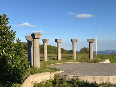 A collection of ancient stone columns stands in a circular formation on a grassy hill under a clear blue sky with scattered white clouds. The columns are weathered and have intricate carvings on their capitals. In the foreground, a circular concrete platform is surrounded by low stone walls and overgrown with grass and wildflowers. A large green tree with vibrant leaves stands to the left of the columns, partially obscuring one of them. In the background, a line of trees and hills can be seen, with a thin white flagpole visible behind the columns. The scene appears serene and quiet, with the bright sunlight suggesting it is daytime, likely late afternoon given the quality of the light. The location appears to be Zemo Nichbisi, Georgia, an area known for its historical sites. There is no visible text in the image.