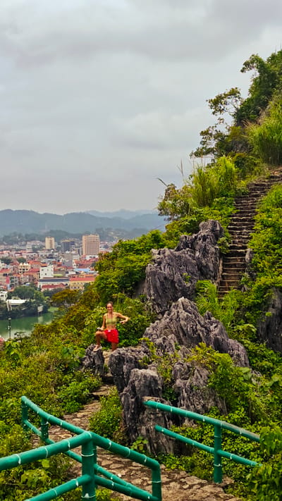 A woman in a green top and red skirt is posing for a photo on a rocky outcrop. She is on a mountain overlooking a city. The city has buildings of various heights and colors, and a body of water. There are lush green trees and plants growing on the mountain. A stone staircase winds its way up the mountain. The sky is cloudy. The woman appears to be in a playful or confident pose. This scene is located in the Tam Thanh Ward of Vietnam.