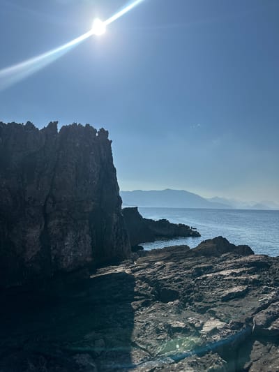 A rugged, dark rock formation dominates the left foreground of this coastal scene in Seoca, Montenegro. The rock face is textured and uneven, with sharp, jagged peaks along its top edge. To the right of the formation, the calm, blue sea stretches towards a distant horizon. The water is clear and reflects the bright sunlight. Faint outlines of layered, hazy mountains are visible in the far distance, appearing as soft silhouettes against the pale blue sky. The sky itself is mostly clear, with the sun positioned in the upper left quadrant, casting a bright glare and lens flare across the image. The rocky ground in the immediate foreground is also dark and uneven, with visible cracks and crevices. Shadows are cast across the rocks, indicating a strong light source. The overall impression is one of a wild and natural coastal landscape.