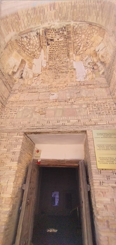 This media file shows an interior view of a historical structure, likely a mosque or madrasah, with intricate brickwork. The walls are adorned with patterned brick ornamentation, some sections of which are worn and chipped, indicating significant age. There is an arched opening on the left side of the image with a lattice-like brick screen.
To the right, a doorway or entrance is visible, framed by wooden beams. In front of the doorway, a red emergency button is mounted on a beige electrical box. Inside the dark opening, a faint, illuminated rectangular shape can be discerned, possibly a display or a distant light source.
A yellow sign with text in Uzbek, Russian, and English is affixed to the wall above the doorway. The text, partially visible, indicates that the structure is the "Magoki Attori Mosque," a "XII-XVI Century" architectural monument "Under State Protection." The lighting suggests an indoor setting, likely during daylight hours, although the interior is dimly lit. The overall impression is one of historical significance and preserved architecture.