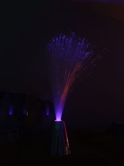 The image shows a fiber optic lamp in a dark environment. The lamp is cone-shaped and the base appears to be silver or reflective. The fibers emerging from the top are illuminated with purple and orange light. In the background, there is a blurry shape that might be another object or a reflection. The lighting is dim, suggesting the lamp is the primary light source. The image does not contain any people, actions, or interactions, and does not provide information regarding the city of Cherkovitsa, Bulgaria.