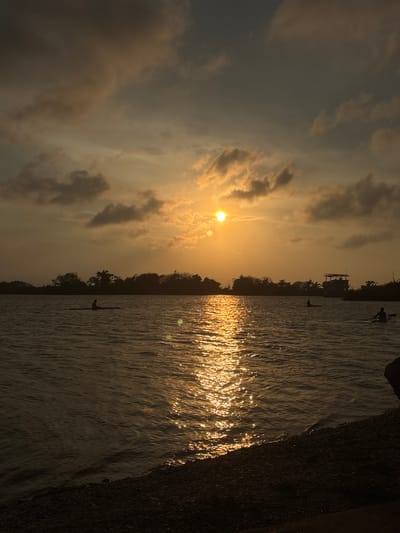 The image depicts a sunset over a body of water, likely a lake or river, in Tampico, Mexico. The sun is low on the horizon, casting a golden glow across the sky and reflecting intensely off the water's surface. There are clouds in the sky, some of which are illuminated by the setting sun. A dark line of trees and foliage can be seen along the horizon. There are people visible in the water, with one person standing and another rowing a narrow boat, possibly a scull. In the upper portion of the scene, a dark structure is visible atop a dense line of vegetation, appearing to be a small building or platform. The foreground shows a dark, textured edge of what could be a wall or shoreline. The entire scene is bathed in a warm, golden light.