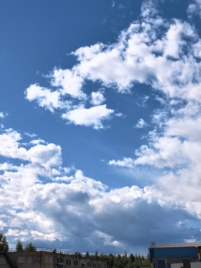 The image shows a bright blue sky filled with fluffy white cumulus clouds. Sunlight filters through the clouds, creating streaks of light in the sky. In the lower part of the image, the tops of buildings and trees are visible, suggesting an urban or industrial setting. The buildings appear to be made of brick and have a somewhat weathered appearance. There is a fence with railings on top of one of the buildings. The overall impression is of a pleasant day with a dynamic sky, likely late morning or early afternoon given the brightness of the sun. The location is identified as Ivanovo, Russia. No people are visible in the image.