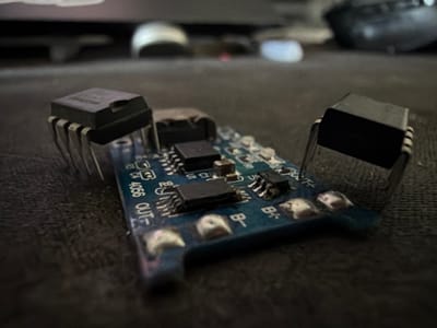 The image is a close-up, shallow depth-of-field photograph of a blue printed circuit board (PCB) with several electronic components. Two integrated circuits (ICs) are prominently visible, one on the left and another on the right, each with multiple pins. Smaller surface-mount components, such as resistors and what appear to be other ICs, are also present on the PCB. Text and markings, including "4056" and "OUT-", are visible on the board. The PCB is resting on a dark, textured surface, possibly a tablecloth or a desk. The background is blurred, showing indistinct shapes that suggest a domestic or workshop environment. There are no people or animals in the image. The image does not depict any elements that would allow for its location to be identified as Torremolinos, Spain.