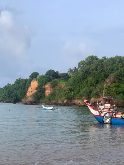 In Mirissa, Sri Lanka, a coastal scene unfolds with a large, colorful fishing boat dominating the right side of the frame. The boat, adorned with a painted image of a woman and intricate patterns, features a white cabin structure and is occupied by two individuals. One man, wearing a white shirt, sits towards the front of the boat, while another man, in a green cap and blue shorts, is positioned further back. A smaller, white and blue boat is anchored in the tranquil water to the left of the larger vessel. Behind the boats, a lush green cliff rises, revealing patches of orange-red rock formations. The top of the cliff is densely covered with trees and vegetation, with a few buildings partially visible amidst the greenery. The sky above is a pale blue, partially obscured by soft, white clouds. The water in the foreground exhibits gentle ripples, with a hint of a sandy shore and the edge of a wave. The overall scene suggests a serene coastal environment, likely during the early morning or late afternoon.
