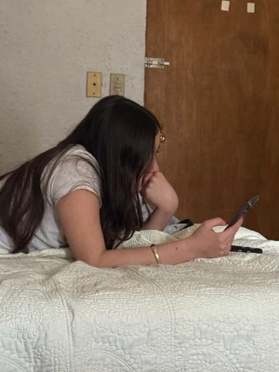 A single female subject with long, dark hair is observed lying prone on a bed, supported by her forearms. She wears a light-colored short-sleeved t-shirt, gold-rimmed eyeglasses, and a gold-colored bracelet on her left wrist. Her right hand, with red-painted fingernails, holds a dark-colored smartphone, towards which her gaze is directed. A dark-colored remote control rests on the light-colored, quilted bedspread beside the smartphone. The bedspread features a repeating circular motif. The immediate background on the left consists of a light-colored, textured wall with two aged electrical wall fixtures: one single outlet and one switch. To the right, a dark brown wooden door is visible, fitted with a metal latch mechanism. Two small, light-colored square adhesive elements are affixed to the upper right quadrant of the door. The specific location within the city of Santiago, Mexico, cannot be determined from the visual information presented.