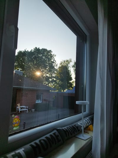 This image is a view from inside a building, looking out a window. The scene outside appears to be a residential area in Schiffdorf, Germany. It is daytime, with the sun visible through the branches of a large tree, suggesting it is either early morning or late afternoon. The weather seems clear, with a bright sky.
The foreground of the image shows a windowsill with several objects. A long, grey cushion with the words "HOME SWEET" printed in a dark, blocky font lies horizontally. Next to it, a small, white, minimalist lamp with a round top stands vertically. A small yellow triangular object and a thin, dark object are also visible on the windowsill. A white curtain is visible on the right side of the window frame, partially obscuring the view.
Outside the window, a brick house with a dark tiled roof is visible. The sun's rays are filtering through the leaves of a large deciduous tree, casting a dappled light. In the yard, there is a white bench and some greenery. A black metal fence runs along the property. A colorful, cartoonish figure, resembling a chick wearing a hat and bow tie, is placed on the ground near the fence.
The overall impression is one of a quiet, domestic scene, possibly a suburban or rural setting. The sunlight creates a warm and peaceful atmosphere.