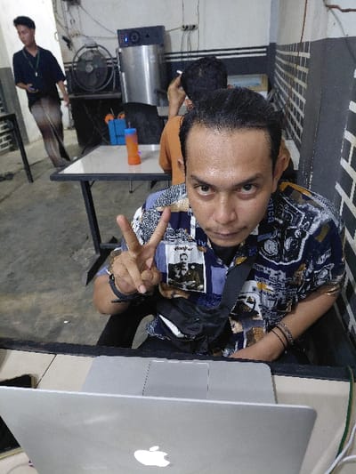 An adult male is seated in the foreground of an indoor location in Mns Alue, Indonesia, making a "V" sign with his right hand and looking directly at the camera with an intense expression. He has dark hair, a sheen on his forehead, and wears a patterned short-sleeved collared shirt adorned with a badge depicting two men. A black cross-body bag is slung over his shoulder, and black wristbands are visible on his left wrist. An open silver Apple laptop rests on a dark-framed table in front of him. In the middle ground, another male, viewed from behind, wears an orange t-shirt and appears to be smoking a cigarette while seated at a similar table. An orange bottle and a blue container are on this table. To the far left, a third male stands, dressed in a dark shirt and light-colored trousers, holding a dark object. The background consists of light-colored walls, partially featuring a dark brick-like pattern and horizontal stripes, with visible electrical wiring. A large dark standing fan and a tall, metallic appliance with blue indicator lights are against the back wall. The floor is dark.