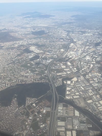 An aerial view of Gebze, Turkey, depicts a sprawling urban and industrial environment under a light hazy sky. A prominent multi-lane highway, identified as a segment of the O-4/E80, bisects the image diagonally, characterized by complex interchanges and visible vehicle traffic. To the left of the highway, dense residential zones consist of numerous light-colored, multi-story buildings. To the right, extensive industrial and commercial parks feature large, rectangular structures with light-colored roofs. In the upper-central distance, Sabiha Gökçen International Airport is visible, identifiable by its runways and terminal infrastructure. Irregular patches of dark green vegetation are interspersed throughout the developed areas. Individual human figures or specific interactions are not discernible.
