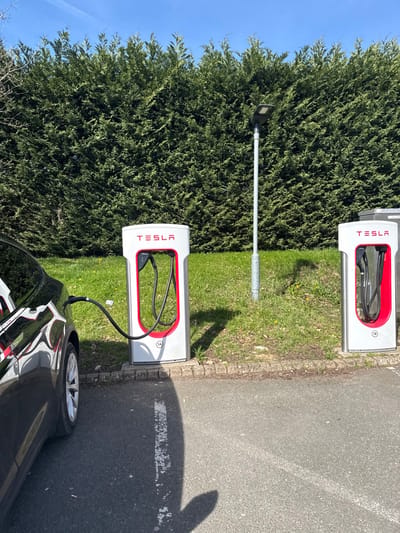 This image depicts an outdoor electric vehicle charging station located in East Hampshire, United Kingdom. The scene is set under a clear blue sky, with bright sunlight casting distinct shadows. In the foreground, asphalt pavement with a white parking line is visible. This pavement transitions to a concrete curb, bordering a grassy verge.
Two white and silver Tesla Supercharger pedestals stand on the grassy verge. The pedestal on the left, labeled "1A," is actively connected to a dark-colored Tesla vehicle. A black charging cable extends from this station into the vehicle's charging port, indicating an active charging session. The rear quarter and a wheel of the dark vehicle are visible on the left side of the frame, its surface reflecting the surroundings. The pedestal on the right, labeled "1B," is unoccupied, with its black charging cable neatly coiled within its red-framed port.
Between the two charging stations, a tall, grey metal pole with a dark, modern LED street light fixture at its top rises from the grass. The grassy verge exhibits patches of exposed soil and a few small yellow wildflowers. A dense, tall, green hedge forms a continuous backdrop across the entire width of the image, separating the charging area from what appears to be an adjacent property or natural area. No individuals are present in the scene.