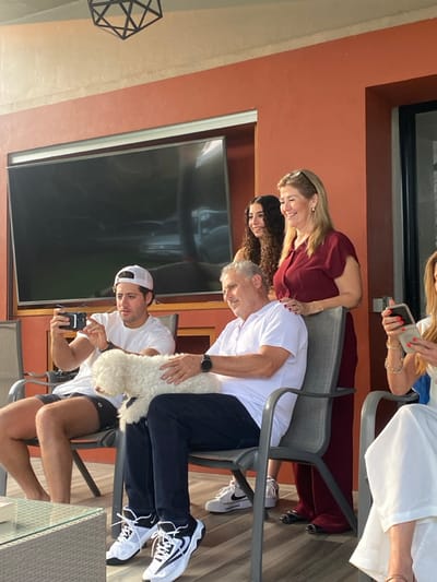 A group of people are enjoying an outdoor gathering. Two women are seated at a table wearing a red and white dress. A younger man with a white cap is holding a phone and taking a picture while an older man is holding a small white dog. Three women stand or lean by the seated people. A large television screen is visible in the background.