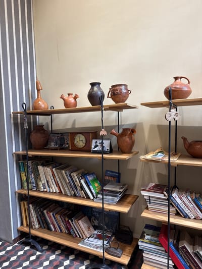 The image displays a three-tiered wooden shelf unit filled with books and decorative ceramic pottery. The shelf unit is positioned against a plain, light-colored wall, with a vertical striped wallpaper visible on the left side. The floor features a patterned tile design with alternating dark red and black diamond shapes.

On the top shelf, from left to right, are several ceramic items: a tall, curved wooden object resembling a flute or a stylized bird, a small reddish-brown pitcher with two spouts, a dark brown ceramic vase with intricate patterns, a reddish-brown pot with carved animal figures, and a reddish-brown pitcher with a handle and decorative lines.

The middle shelf contains more ceramic pieces. On the left, there's a large, reddish-brown jug with a rounded body and a narrow neck. Next to it are several framed photographs or small paintings, including a clock with a circular face and Roman numerals. To the right, there is another reddish-brown jug with two spouts, and a small, reddish-brown teapot with a spout and handle.

The bottom shelf is densely packed with books of various sizes and colors, some standing upright and others leaning. The spines of the books show titles in different languages, including English and possibly Armenian.

The overall scene suggests a display within a shop or a home, possibly a museum or a souvenir shop, given the presence of artisanal pottery and books. The location is identified as Dilijan, Armenia.
