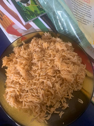 The image shows a plate of cooked rice. The rice is light brown and appears to be seasoned. The plate is made of a clear material with a gold rim. The rice is positioned centrally on the plate.
To the left of the plate are books, including one titled "Mathematics" and another with "Physics Writers Series Creatic" visible. There is also a package that is labeled in part "...better? ...we source our maize carefully...". The background is a dark blue or purple color.