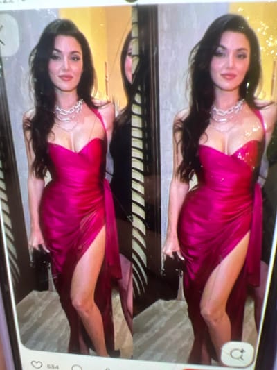 The image displays a woman in a vibrant magenta dress with a high slit, posing in what appears to be an indoor setting with a reflective surface creating a mirrored effect. She has long, dark hair and is wearing a chunky silver necklace. The background is softly lit and somewhat indistinct, with hints of textured walls and possibly decorative elements. The overall impression is of a fashion or event setting. There is no discernible connection to the city of Kara-Dıykan, Kyrgyzstan, as the image provides no visual cues or context to suggest this location.
