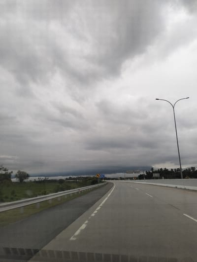 The image depicts a highway in Baksukon, Indonesia, under a heavily overcast sky. The road curves to the left, with multiple lanes visible. A metal guardrail runs along the left side of the road, separating it from a grassy area and trees. Beyond the grass, a distant treeline of palm trees suggests a tropical location. A tall lamppost with two lights stands on the right side of the road. Several road signs are visible, including a yellow directional sign with a curved arrow and another larger overhead sign with text that is illegible due to distance and resolution. The clouds are dark and dense, indicating impending rain or a storm. The overall mood is somber and somewhat dramatic due to the weather. The time of day is difficult to ascertain precisely due to the cloud cover, but the lighting suggests it is daytime. There are no people visible in the image. The scene captures a moment of travel on a highway, with the natural landscape contrasted by the man-made road infrastructure.