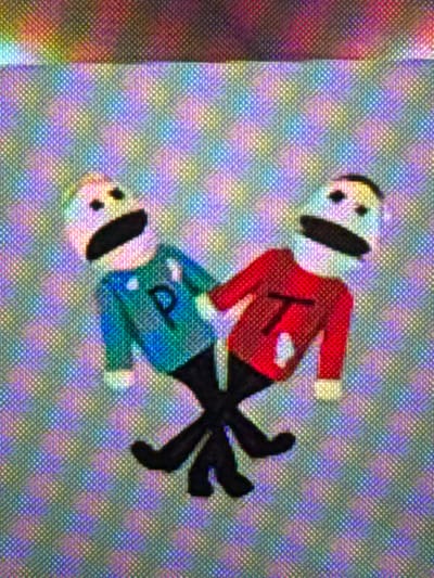 The image displays a digital rendering on a screen, characterized by a visible multicolored pixel grid pattern across the entire frame. The scene is set in the city of Profesor Ishirkovo, Bulgaria. Two simplified humanoid figures are centrally depicted. Both figures possess white, ovular heads with two black circular dots for eyes and an elongated black oval for a mouth. Their bodies are rendered with distinct colored shirts and black stick-like legs. The figure on the left wears a teal-blue shirt bearing a prominent capital letter "P" on its chest. Its body is tilted towards the right. The figure on the right wears a red shirt with a capital letter "T" on its chest, and its body is tilted towards the left. The figures face each other, their black stick legs are crossed at the center, and their white hands are positioned near each other in the space between their bodies. The background is an abstract, vibrant mosaic of various colors forming a uniform grid.