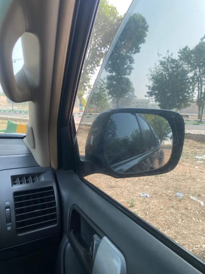 This image depicts a static scene captured from the interior of a vehicle, looking out the passenger-side window, located in Yan Dala, Nigeria. The foreground shows internal vehicle components: a beige A-pillar on the left, a black dashboard section with two sets of horizontal air vents, and a black door panel with a silver door handle. A tinted side window is partially rolled down. The exterior view reveals a black side mirror, reflecting part of the vehicle's body and the environment behind. The ground outside the vehicle consists of dry, sparse vegetation and dirt, with several pieces of white litter (likely plastic) scattered on its surface. In the mid-ground, a paved area or road with light-colored curbs and green and yellow markers is visible. The background is dominated by multiple trees with green foliage under a light, clear sky. A building structure is faintly discernible in the far distance, behind the trees. No individuals are present in the scene, and no actions or interactions are depicted.
