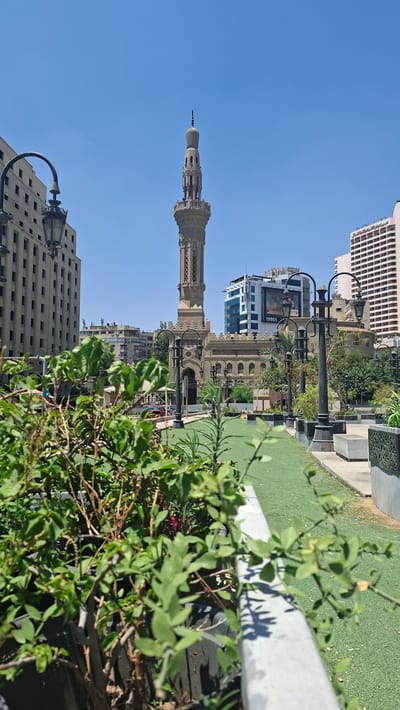 A tall, ornate minaret, characteristic of Islamic architecture, dominates the frame, reaching towards a clear, bright blue sky. The minaret, made of a light-colored stone, features intricate carvings and balconies, with a decorative finial at its apex. The image appears to be taken on a sunny day, likely in the late morning or early afternoon given the bright sunlight and distinct shadows.
In the foreground, lush green foliage, possibly from bushes or trees, partially obscures the view. Beyond the greenery, a well-maintained public space is visible, with neatly trimmed grass and a row of decorative black lampposts. The lampposts have a classic design with curved arms.
Further in the background, a mix of architectural styles is evident. There is a building with a repeating pattern of windows, suggesting a modern or neoclassical design. Adjacent to it, another structure showcases more traditional Islamic architectural elements, possibly a mosque or historical building, with arched windows and decorative facades. A modern high-rise building with glass panels is also present, and a billboard displays the year "1985" and some unreadable text.
The scene suggests an urban environment in Cairo, Egypt, likely a public square or park area, where historical and modern structures coexist. The overall mood is serene and bright, showcasing a blend of culture, history, and urban life under a clear sky. There are no people visible in the frame, focusing the attention on the architecture and the public space.