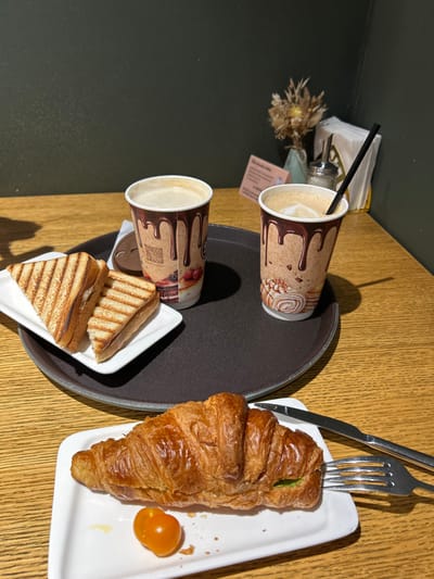 The scene is an indoor cafe setting in Voskresensk, Russia, presented on a light brown wooden table. A dark brown circular tray occupies the midground, holding two disposable paper cups patterned with chocolate drips. Both cups contain a light brown beverage, likely coffee; one cup has a black straw inserted, and a separate brown cup lid rests near the other cup. Adjacent on the tray, a white rectangular plate holds two triangular grilled sandwiches with visible grill marks. In the foreground, a second white rectangular plate features a golden-brown croissant with a green filling visible at one end, alongside a single orange cherry tomato. A silver fork is partially embedded in the croissant, and a silver knife lies parallel to it on the plate. The background consists of a dark green wall. Tabletop decor behind the tray includes a small vase containing dried flowers or botanicals, a silver sugar dispenser, a white napkin holder with white paper napkins, and a small, light-colored rectangular informational card. No individuals are visible.