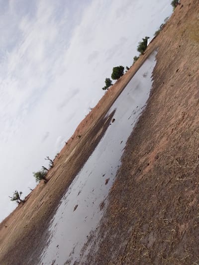 The image shows a landscape in what appears to be the Mulke Barikin Sayejo region of Nigeria. The sky is overcast with a cloudy sky. The ground consists of reddish-brown soil. A body of water, possibly a puddle or shallow stream, reflects the sky and cuts through the landscape. Sparse vegetation and scattered trees appear on the horizon and hillside. The ground near the water's edge appears muddy with scattered dry grass or straw. The image is tilted.