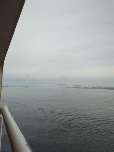 This is a photograph taken from a vessel on the water, looking towards a city skyline and a large suspension bridge. The sky is overcast and a dull grey. The water in the foreground is choppy and dark grey. On the left side of the frame, a white railing and part of the vessel's structure are visible.
In the distance, a prominent cable-stayed bridge spans the water, with two tall pylons supporting its deck. The bridge appears to be a significant piece of infrastructure. Beyond the bridge, a city skyline with various buildings, including some industrial structures like chimneys, can be seen. This scene is likely of the Yokohama Bay Bridge or a similar bridge in Yokohama, Japan, given its distinctive architecture and the presence of a port. There are no people visible in the image.