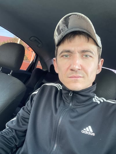 The media file is a selfie of a person in a car, likely taken during the daytime. The person is wearing a black Adidas tracksuit with white stripes on the sleeves and a baseball cap with a newspaper print design. Their green eyes are looking directly at the camera. The background shows the interior of the car, including car seats and the roof lining. Outside the car window, buildings and trees are visible, suggesting an outdoor setting. There is a visible Adidas logo on the tracksuit. The image is a close-up of the person's face and upper body, rotated 90 degrees counterclockwise.