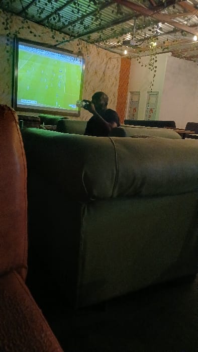 A blurry, tilted shot shows a man pouring a drink in front of a large, illuminated screen displaying a green soccer field. Metal latticework covered in greenery adorns the wall to the left, while a tan wall with windows stretches across the top. A large, green, heavily textured sofa dominates the right side of the frame, obscuring the view and adding to the sense of cramped space. A brown leather sofa extends across the bottom of the frame.