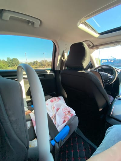 The image captures the interior of a vehicle, likely a car, from a rear passenger perspective. Sunlight streams in through the open sunroof, illuminating the cabin. The focus is on a baby car seat in the foreground, draped with a pink and white blanket featuring small red strawberry patterns and cartoon animal figures. A baby's arm is visible, peeking out from under the blanket.

Behind the car seat, the back of a driver's seat is visible, occupied by a person wearing a dark shirt. The steering wheel and dashboard of the car are partially in view, with the Ford logo visible on the steering wheel. The car's interior appears to be clean and well-maintained.

Outside the car windows, a clear blue sky suggests a sunny day. Green trees and some light posts are visible in the distance, indicative of an outdoor environment. The presence of a car and its interior elements suggest the scene is set in a location with roads and possibly a parking area. The location being Herasymivka, Ukraine, implies this scene is taking place within that geographical context, though specific landmarks to pinpoint the exact location within the city are not evident from the image alone.