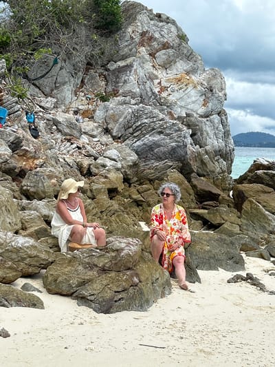 Two women are sitting on rocks on a sandy beach in Phru Nai, Thailand. The woman on the left is wearing a straw hat and a white dress. The woman on the right is wearing a floral print kimono. In the background, there is a large rock formation and the ocean. The sky is cloudy. The scene appears to be a casual outing at a beach location.