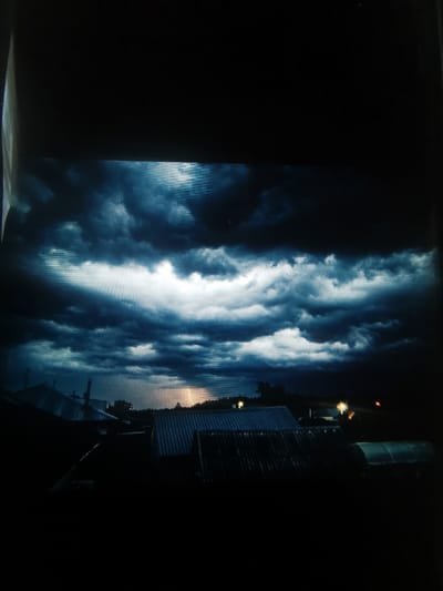 The image depicts a dramatic, stormy sky over a rural landscape. Dark, turbulent storm clouds dominate the upper portion of the frame, with breaks of lighter clouds and a visible patch of sunlight or lightning in the distance. Below the sky, the silhouettes of several rooftops are visible, suggesting a small settlement or village. Some of these structures appear to have corrugated metal roofs. In the far distance, hints of trees and possibly some lights suggest the presence of further habitation.
The time of day is indeterminate due to the storm, but the lighting suggests either dusk or a very overcast day. The weather is clearly stormy, with the dark clouds and potential lightning indicating a significant weather event. The overall mood is intense and foreboding. No people are visible, but the presence of buildings implies human activity in the area. There is no visible text in the image. The landscape suggests a rural setting, potentially in a northern climate given the hint of a greenhouse-like structure visible on the far right.