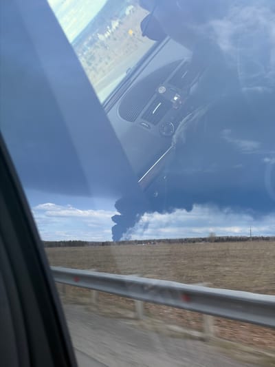 The image shows a view from inside a moving vehicle, likely a car, traveling along a road. A guardrail is visible along the right side of the road, with a field or open area beyond it. The dashboard of the vehicle is partially visible, with vents and controls. The windshield reflects the sky and clouds. The city of Yugo-Kamskiy, Russia is not discernible from the provided image.