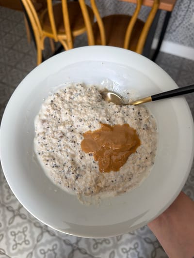 A close-up overhead shot shows a white bowl filled with oatmeal and topped with a dollop of peanut butter. A spoon with a black handle and gold tip rests in the oatmeal. The oatmeal appears to contain chia seeds and is mixed with a white liquid, likely milk or yogurt. The bowl is placed on a table with a patterned grey and white tile floor. In the background, the blurry image of a wooden chair with a light-colored seat is visible. A person's hand and arm are visible on the right side of the image, reaching towards the bowl. The lighting suggests it might be daytime. The setting appears to be indoors, possibly a dining area or kitchen. There is no visible text in the image.