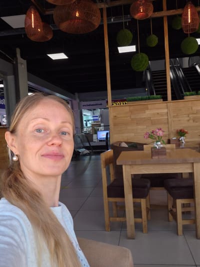 A blonde woman with blue eyes and freckles, wearing a white t-shirt and a pearl earring, takes a selfie in an indoor setting. She has long, light brown hair. Behind her, to the left, are dark ceilings with decorative pendant lights and some green spherical decorations. To the right, there's a wooden wall paneling, a wooden table with chairs, and a vase of pink flowers. In the background, escalators are visible. The floor appears to be tiled. The sign in the background reads "ARSE'N'IEV" indicating the location is Arseniev, Russia.