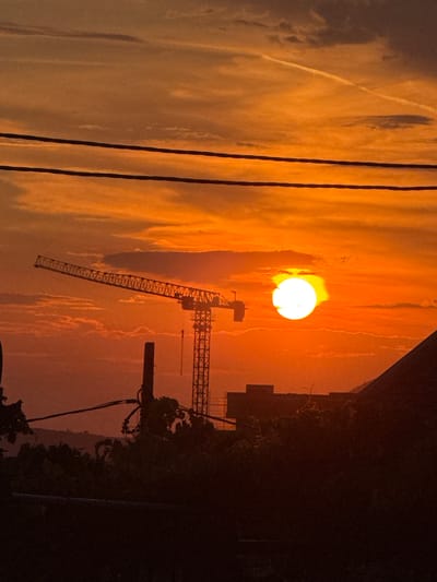 This image captures a vibrant sunset over Burtaiši, Montenegro. The sky is dominated by intense shades of orange and red, with wispy clouds and a contrail adding texture. A large, bright sun is prominently featured, appearing as a glowing orb. In the foreground, silhouetted against the colorful sky, are several elements: power lines stretch across the upper portion of the frame, a construction crane with its boom extended is visible on the left, and a series of dark, silhouetted structures and vegetation occupy the lower part of the image. One of these structures appears to be a building, and a sloped rooftop is visible on the far right. The overall scene suggests a developing urban or industrial area undergoing construction, juxtaposed with the natural beauty of the sunset.