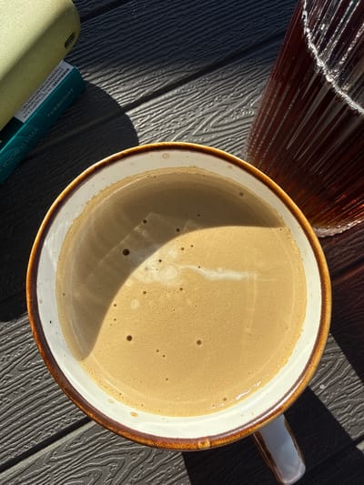 A top-down shot shows a mug filled with a light brown beverage, likely coffee with cream. The mug has a brown rim and a white interior. A glass carafe, partially visible in the upper left corner, contains a dark liquid. A green and blue package labelled "IQOS ILUMA" and "TURQUOISE" is visible at the bottom left. The image is taken in bright sunlight, casting a shadow across the mug and the underlying surface, which appears to be dark wood. The text on the packaging is written in Bulgarian, suggesting the image was taken in Bulgaria. There are no people visible. It is not possible to identify the location as Sandanski, Bulgaria based on the visual information alone.