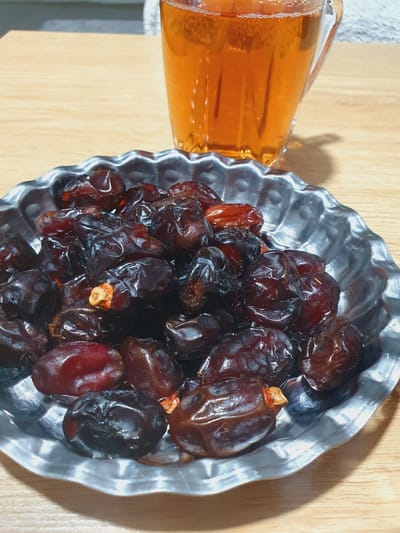 A close-up, overhead shot shows a metallic, fluted dish overflowing with dark, plump dates. The dates are a deep, rich brown, some with a reddish hue, and appear glistening, suggesting freshness or a light coating of moisture. A few of the dates reveal small, lighter-colored remnants of their pits. Behind the dish, a glass mug filled with a golden-amber liquid, likely tea, is visible. The mug has vertical ridges and a slight foamy surface at the top. Both the dish and mug rest on a light-colored wooden surface, likely a table. The background is softly blurred, with hints of a light-colored fabric or surface and a subtle shadow cast by the mug. The scene is lit, creating reflections on the dates and the mug. The image is likely a still life, capturing a common refreshment. There is no indication of location in Kheyrabad, Iran, or any people or actions taking place.
