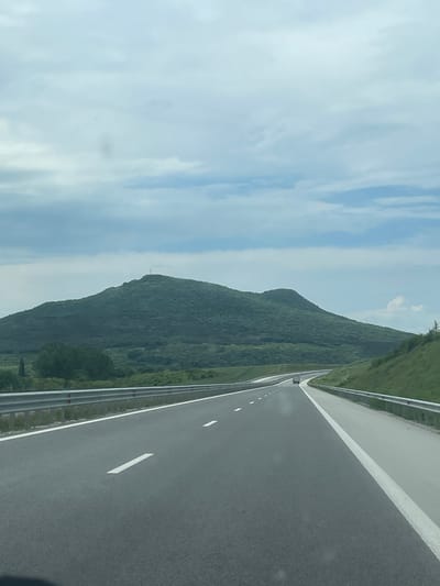 The image shows a highway in Belokopitovo, Bulgaria. A dense green mountain range rises along the left side of the road. The road itself is divided by white lines and features a metallic guardrail along the shoulder. The sky is partly cloudy.