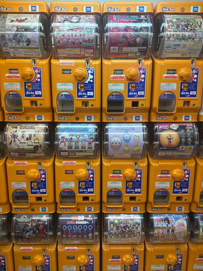 This image depicts a wall densely packed with Gashapon (capsule toy) vending machines within a commercial establishment in the city of Nakano, Japan. The scene displays at least five rows and six columns of uniformly designed machines, primarily in a bright yellow-orange color. Each machine features a clear plastic dome at the top, showcasing various encapsulated collectible toys, figures, and keychains.

Essential details include:
- **Machines:** Each unit is equipped with a coin slot for Japanese Yen (prices like ¥300, ¥400, ¥500 are visible), a large yellow circular knob for dispensing, and a rectangular chute for capsule retrieval. Blue instructional labels and "Gacha." branding are prominent.
- **Contents:** The transparent domes display a wide array of miniature collectibles from various franchises, including "MOBILE SUIT ENSEMBLE" (Gundam-related), "SANDLAND", "JOJO'S BIZARRE ADVENTURE", and numerous character-based figures and accessories, indicative of popular Japanese anime, manga, and game culture.
- **Branding:** "TAKARA TOMY ARTS" and "BANDAI" logos are visible on some machines, identifying manufacturers.
- **Environment:** The overall arrangement suggests an indoor retail or arcade setting dedicated to these automated toy dispensers. No people are visible within the frame.