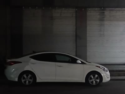 A white sedan is parked in front of a closed garage door with horizontal slats. The car is facing towards the right of the frame. The scene is dimly lit, suggesting it is nighttime or indoors. The garage door appears to be made of metal and has some visible markings or smudges. The car's headlights and taillights are not illuminated. The overall environment is urban, possibly a commercial area in Vagharshapat, Armenia, given the context. There are no people visible in the image. The car's wheels are silver alloy rims with five spokes. The vehicle's bodywork is clean, and the windows are tinted.