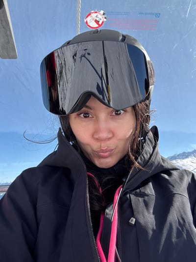A person is depicted in a close-up, selfie-style image, situated within what appears to be a ski lift cabin in St. Christoph, Austria. The individual, whose gender is not explicitly stated but presents as female, wears a black ski helmet with large, dark mirrored goggles positioned on top of the helmet, exposing her eyes. She is dressed in a black zippered ski jacket, under which a bright pink layer and a dark base layer are visible. Her mouth is puckered in a "kissing" or playful expression. Above the person's head, a metallic cable and a transparent panel with a red and white circular warning sticker (depicting a crossed-out figure) are visible, along with faint text including "Doppelmayr," identifying the lift manufacturer. The background consists of a clear blue sky and distant snow-covered mountains, indicating an alpine environment. Reflections within the goggles show a snowy slope and elements of a ski lift structure. The scene is illuminated by bright daylight.