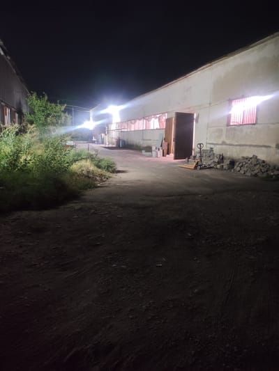 The image depicts an outdoor scene at night in Dangir, Uzbekistan, likely in an industrial or storage area. The foreground shows a dark, uneven dirt or gravel path. To the left, overgrown green bushes and vegetation are prominent. In the mid-ground, a long, light-colored building with several windows is visible, some of which are illuminated from within. One large, dark doorway is open, revealing some boxes or materials inside. Adjacent to the doorway, a yellow pallet jack is parked next to a pile of rough stones. Further along the building, more piles of stones are scattered. The background is dark, indicating the night sky, with bright artificial lights casting beams from the building, creating lens flares. Another building, with a corrugated metal exterior, is partially visible on the far left. The scene appears quiet and still, with no people visible.