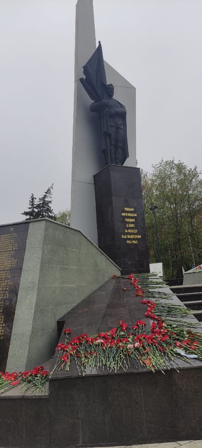 This is a photograph of a war memorial located in the city of Fryazino, Russia. The monument includes a statue of a figure draped in what appears to be a flag, positioned atop a gray, angled structure. Adjoining this is a darker, vertically oriented block inscribed with Cyrillic text in gold lettering. The inscription "ВОИНАМ ФРЯЗИНЦАМ, ПАВШИМ В БОЯХ ЗА ПОБЕДУ НАД ФАШИЗМОМ 1941-1945" translates to "To the soldiers of Fryazino who died in battles for victory over fascism 1941-1945". The base of the memorial has an angled section covered in dark stone, bearing an engraved list of names. Red carnations are laid along the base of the memorial, indicating remembrance. The sky is overcast and gray. Trees are visible in the background.
