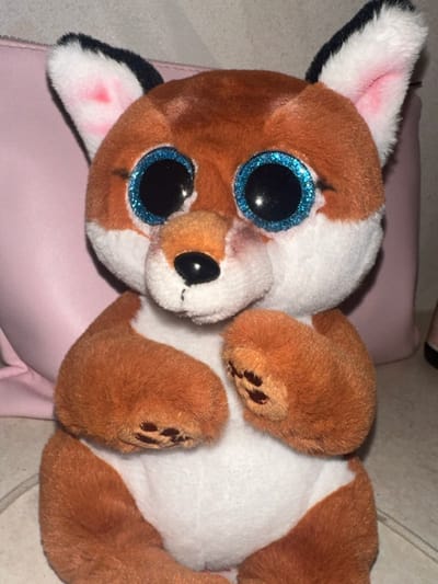 This image, taken in Fowlis Easter, United Kingdom, depicts a plush toy fox positioned centrally, facing directly forward. The toy features an orange-brown and white color scheme. Its large, round eyes consist of black pupils encircled by bright blue glittery irises. A small, black, oval-shaped nose is present on a white muzzle. The ears are white with black tips and pink interior fabric. A faint dark smudge is visible on the right side of the muzzle, near the eye, along with a slight pinkish discoloration below the left eye and on the upper right side of the muzzle. The front paws, which are orange-brown with three stitched dark brown paw pad details, are slightly raised and positioned in front of the white chest. The lower body and legs are orange-brown. The toy is situated on a light-colored, possibly tiled surface. A light pink object, consistent with a bag or pouch, is visible in the background, extending horizontally behind the plush toy.