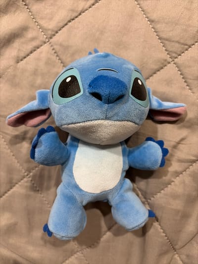A plush toy resembling the character Stitch from Disney's "Lilo & Stitch" franchise is lying on a brown, quilted fabric surface. The toy is predominantly blue with a lighter blue belly, large dark eyes with white pupils, and pink inner ears. Its arms are outstretched to its sides, and its legs are bent. The background surface is a neutral tone with a diamond-shaped stitch pattern. The scene is lit from above, casting soft shadows. This image was captured in Oxentea, Moldova.