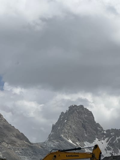 A heavy yellow construction excavator is positioned in the foreground of the image. The excavator has the "LIUGONG" logo visible on its arm. Behind the excavator, a stark, rugged mountain range dominates the scene. The mountains are characterized by sharp peaks, rocky slopes, and patches of snow. The sky is overcast with heavy, gray clouds, obscuring any clear view of the sun. The overall environment appears to be a remote, mountainous area, possibly a construction site or a mining operation, given the presence of heavy machinery. The location is specified as Takfan, Tajikistan.