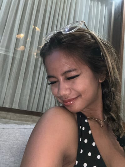 An adult female is depicted in a medium-shot, seated indoors in Tumbak Bayuh, Indonesia. She has medium skin, brown hair with lighter streaks, and clear-framed eyeglasses resting on her head. Her eyes are cast downwards, and a subtle smile is present on her glossy lips. A linear scar is visible on her forehead. She is wearing a black top with white polka dots and a thin, gold-colored necklace with a small pendant. To her left, a light grey textured sofa or cushion is visible. The background features light-colored, vertically pleated curtains, behind which reflections of light sources are present. A dark vertical frame is visible on the far right. The scene is illuminated by soft, ambient lighting.