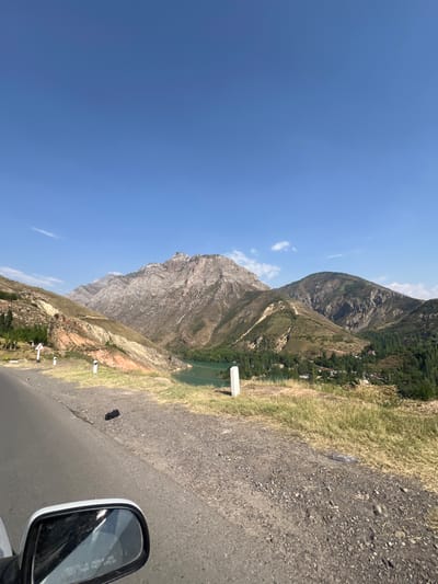 The image captures a scenic view of a mountainous landscape with a body of water and a small village. The foreground shows a paved road with dry grass and gravel on the shoulder. A car's side mirror is visible in the lower left corner, reflecting the sky and some of the surrounding environment. In the mid-ground, a turquoise body of water, likely a reservoir or lake, is nestled between rolling hills. Several white posts, possibly road markers, are placed along the roadside. Further in the background, a majestic mountain range with rugged, rocky peaks dominates the horizon under a clear blue sky with a few wispy clouds. The slopes of the mountains are covered in sparse vegetation, with patches of green trees, especially near the water's edge and around the village. The village itself appears small, with a few scattered houses. The overall impression is one of natural beauty and a sense of remoteness, consistent with the natural surroundings of Brichmulla, Uzbekistan, a region known for its mountainous terrain and reservoirs. There are no people visible in the image, and the objects present are natural elements, road infrastructure, and what appears to be litter (a plastic bottle).