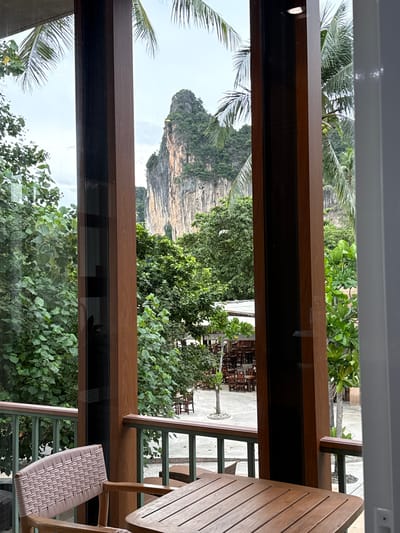 The image captures a view from a balcony overlooking a courtyard with tables and chairs, framed by wooden pillars. In the background, a towering limestone karst mountain, characteristic of the Krabi province, rises dramatically against a cloudy sky. Lush green trees and palm fronds surround the courtyard, suggesting a tropical environment. The foreground features a wicker chair and a wooden table on the balcony. The scene is located in Ban Ao Nang, Thailand, a popular tourist destination known for its stunning natural landscapes.