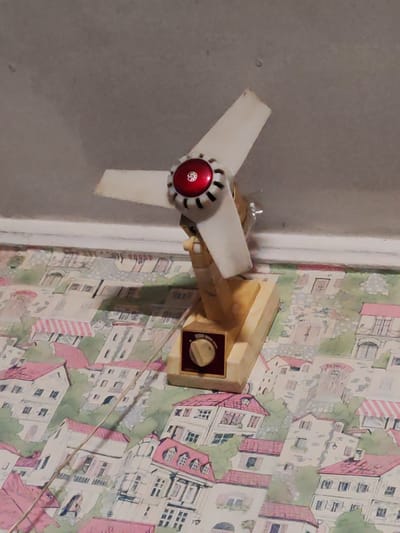The image shows a close-up view of a vintage-style electric fan with white blades and a red and silver center, positioned against a patterned wallpaper that depicts a European-style village with buildings featuring pink roofs. The fan is mounted on a light brown wooden base, which has a dial with a knob on the front. A thin, light-colored wire runs from the fan and drapes across the wallpaper. The background above the wallpaper appears to be a plain, textured wall. The overall impression is of an indoor setting, likely a room, with the fan serving as the primary subject. There is no indication of the fan's current operational status. The location of Kashino, Russia is not depicted or suggested in this image.