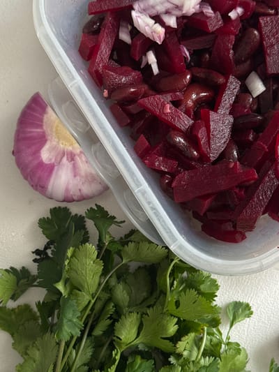 The image shows a plastic container filled with diced beets, kidney beans, and chopped white onion. Next to the container, a half-cut red onion sits alongside a bunch of fresh cilantro. The scene is situated on a plain white surface. There is no evidence to suggest a link to the city of Akulovo or Russia.
